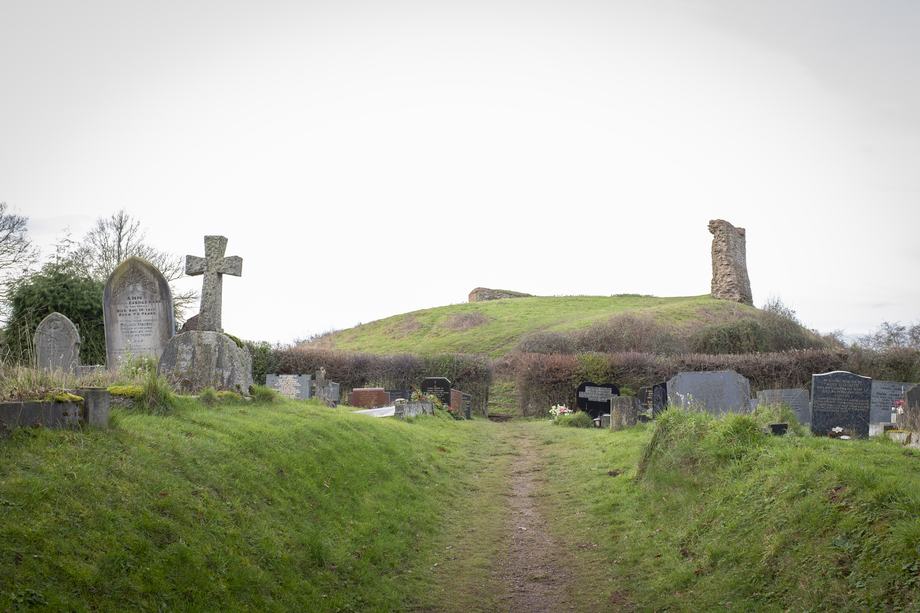 A narrow dirt path through a grassy cemetery lined with weathered headstones and a stone cross leads toward a grassy mound topped by the ruins of an old tower under a pale overcast sky.