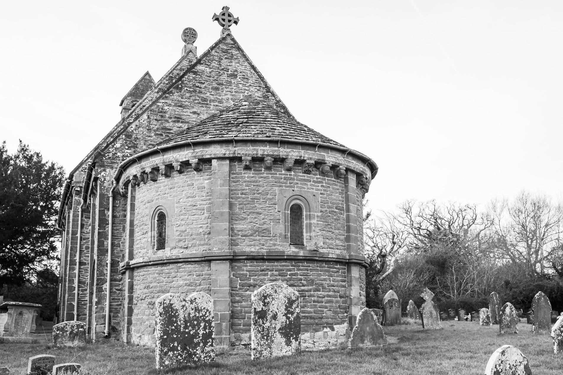 Black-and-white photo of an old stone church with a rounded apse and small arched windows, surrounded by weathered gravestones in a grassy churchyard, bare trees and an overcast sky.