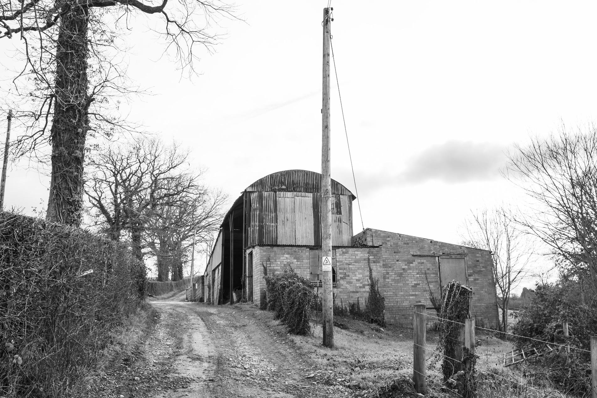 Black-and-white photo of a rural dirt lane curving past a corrugated metal and brick barn, bare winter trees, a lone utility pole and muddy tracks.