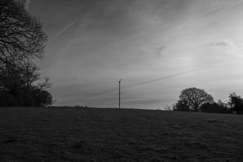 Black-and-white landscape: a gently sloping grassy field topped by a solitary utility pole, framed by bare trees at left and right beneath a wide, cloud-streaked sky with faint airplane contrails.