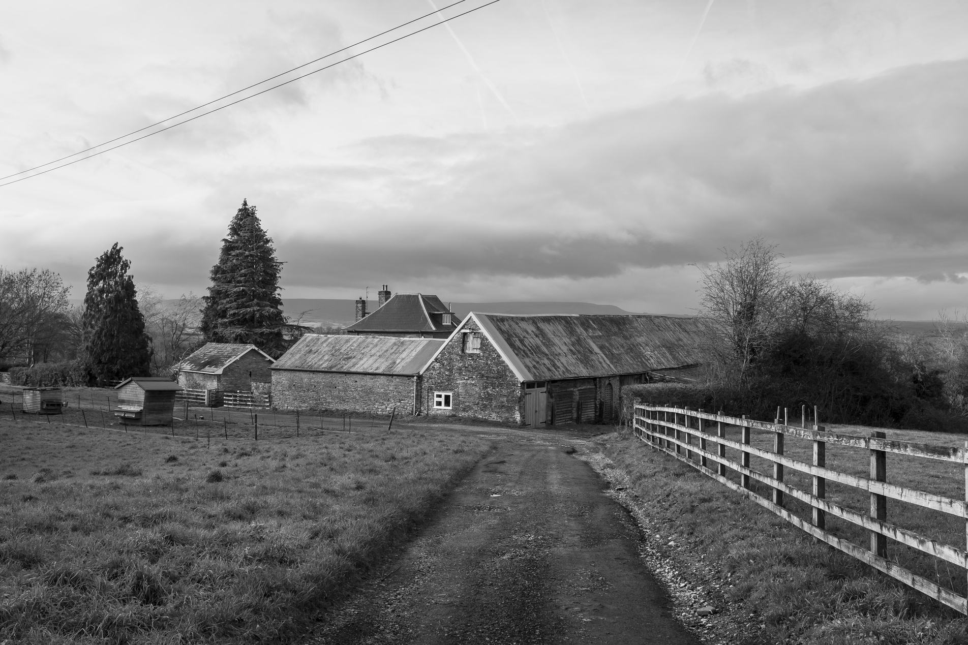 Black-and-white rural scene: a gravel lane curves toward stone barns and outbuildings, a wooden fence lines the right, grassy pasture and tall trees under heavy clouds.