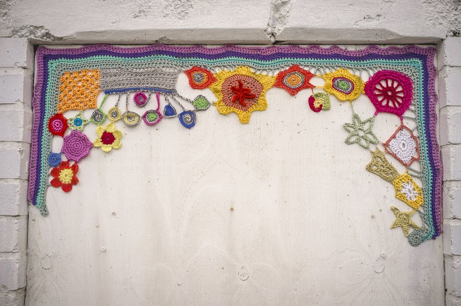 Colorful crocheted yarn border with flowers and geometric motifs draped across the top and upper sides of a white painted wooden panel set in a brick frame.