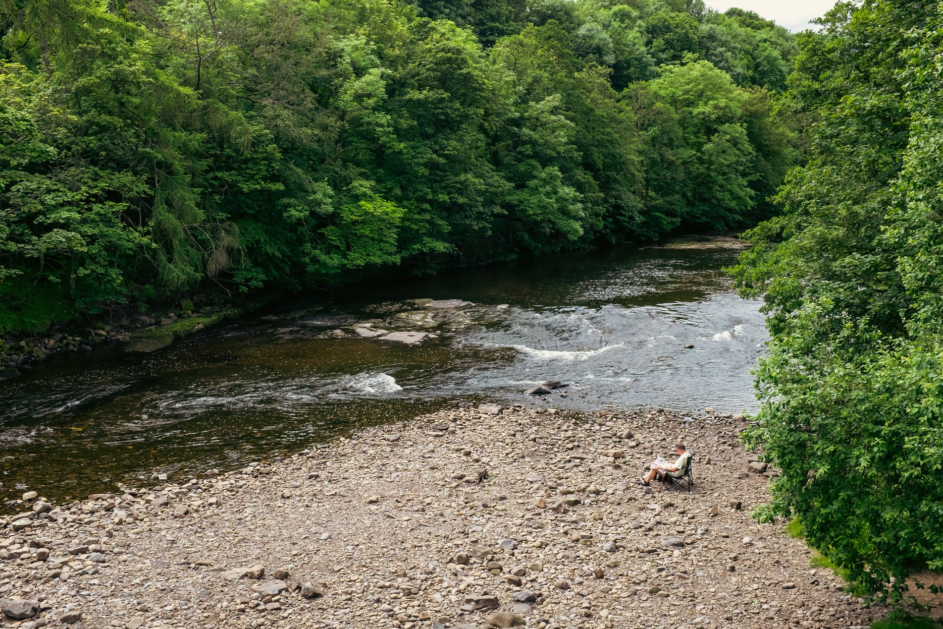 man sitting in deck chair reading a paper alongside the river wear